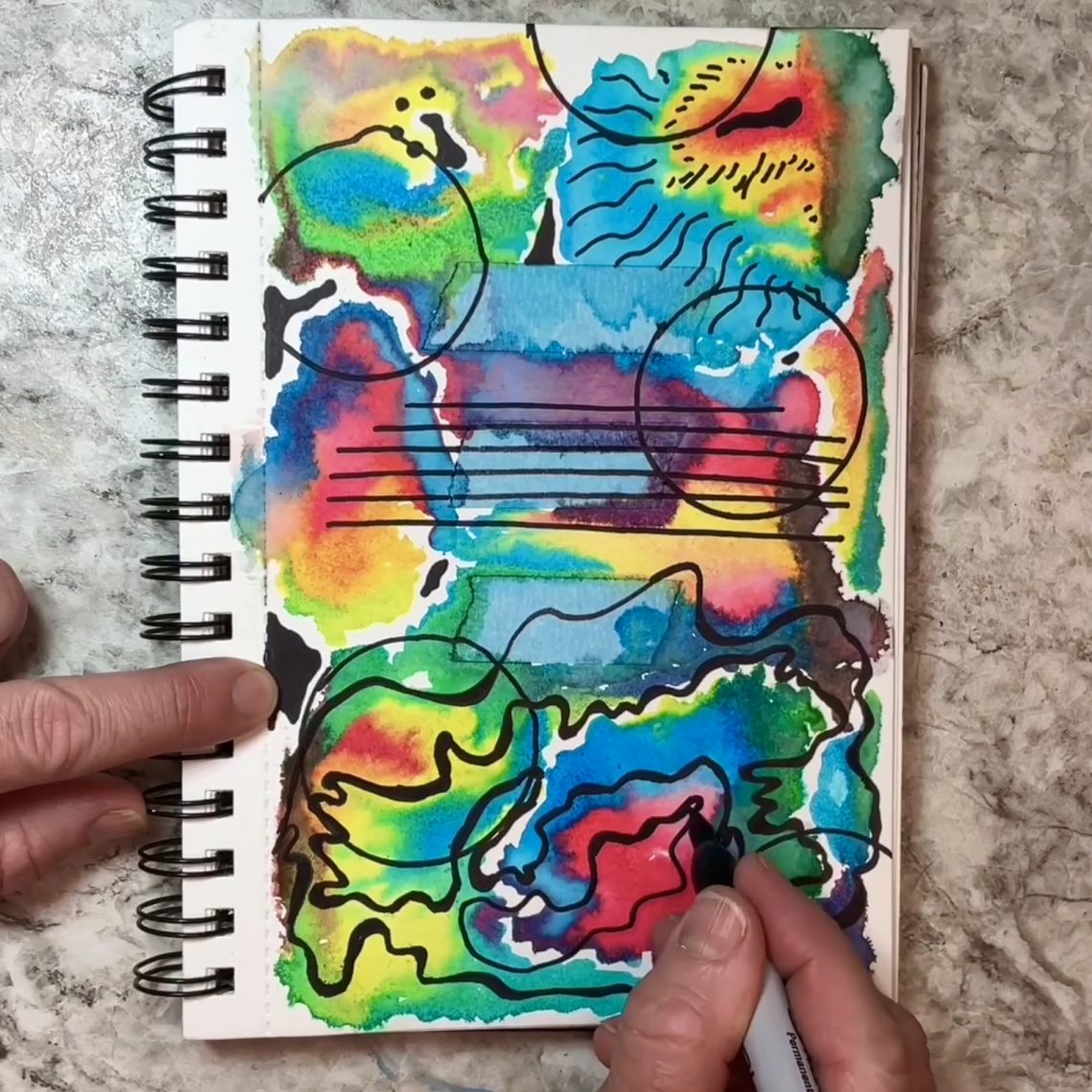 My hand drawing lines and other designs on a background of bright watercolor colors on a 52-Week Art Journal Journey art journal page.